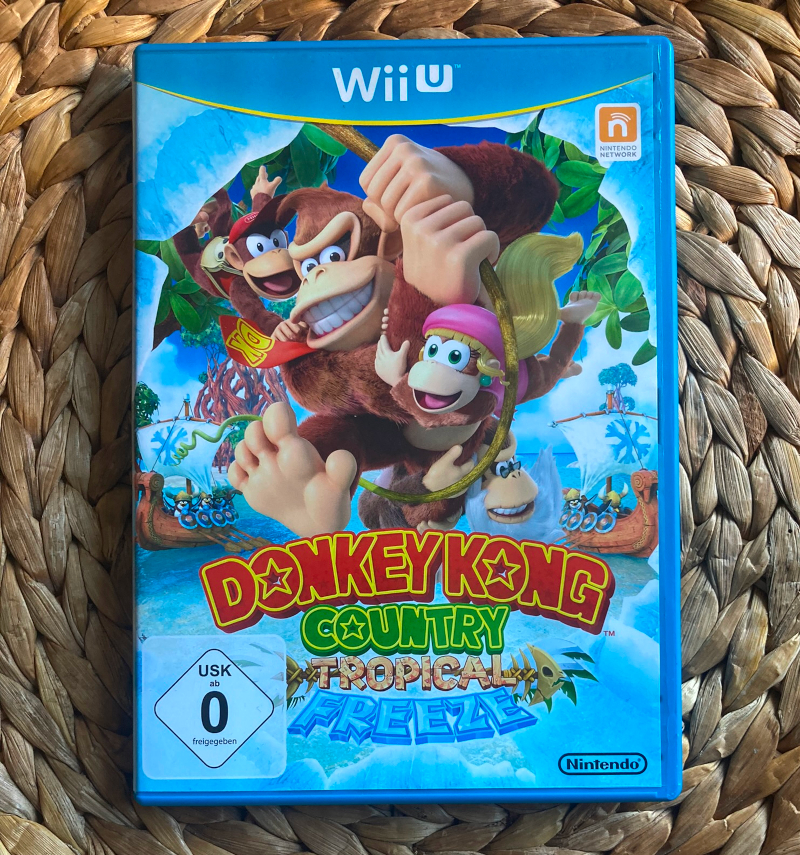 Wii U Spielepackung von Donkey Kong Country: Tropical Freeze. Ein Blick durch ein Eisloch, hinter dem die Kong Familie zu sehen ist, daneben Wikingerpinguine in ihren Schiffen.